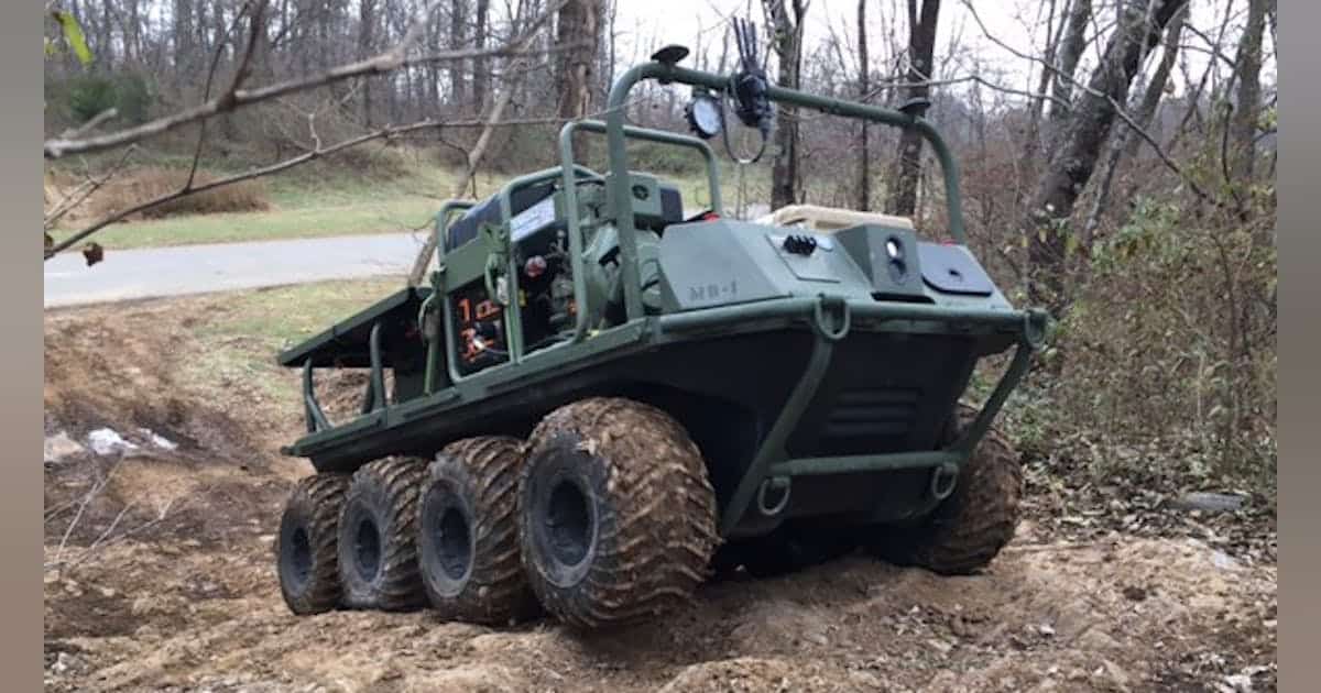 Military-style six‑track all‑terrain vehicle on a dirt trail in a wooded area.