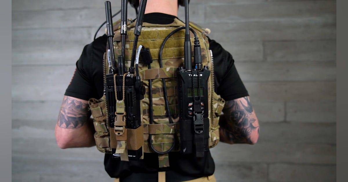 Back view of a person wearing a MOLLE tactical vest loaded with magazines, pouches, and communication gear, with tattoos on the arms and a gray wall background.