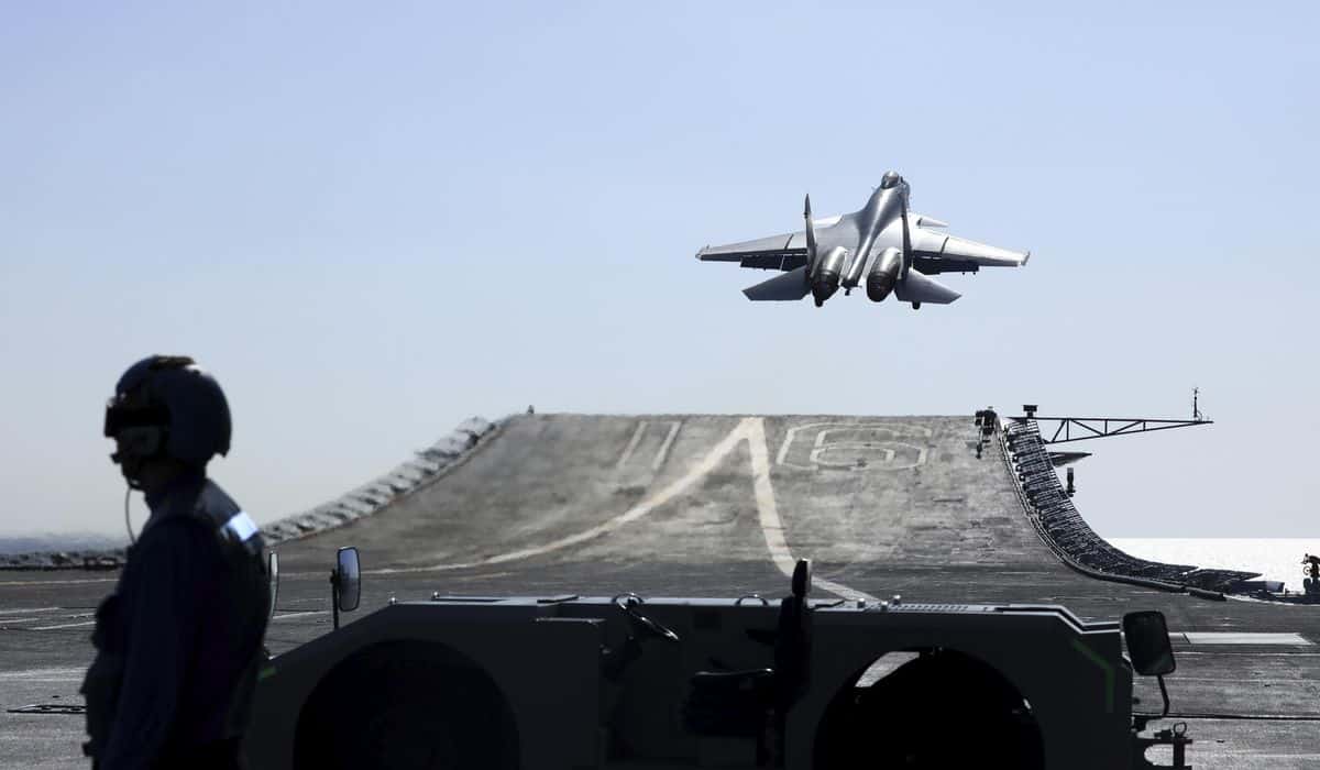 Fighter jet launches off an aircraft carrier catapult, rising above the deck with a crew member visible in the foreground.