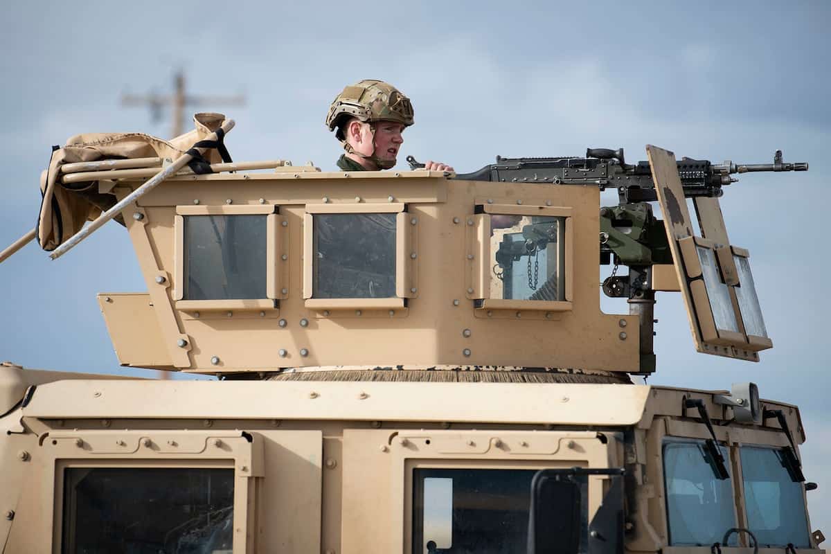 Soldier in helmet peering from the hatch of a tan armored vehicle with a mounted machine gun