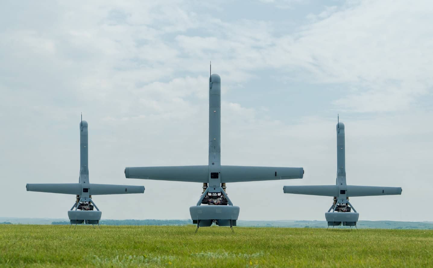 Three large gray unmanned aircraft parked on a grassy airfield under a cloudy sky.