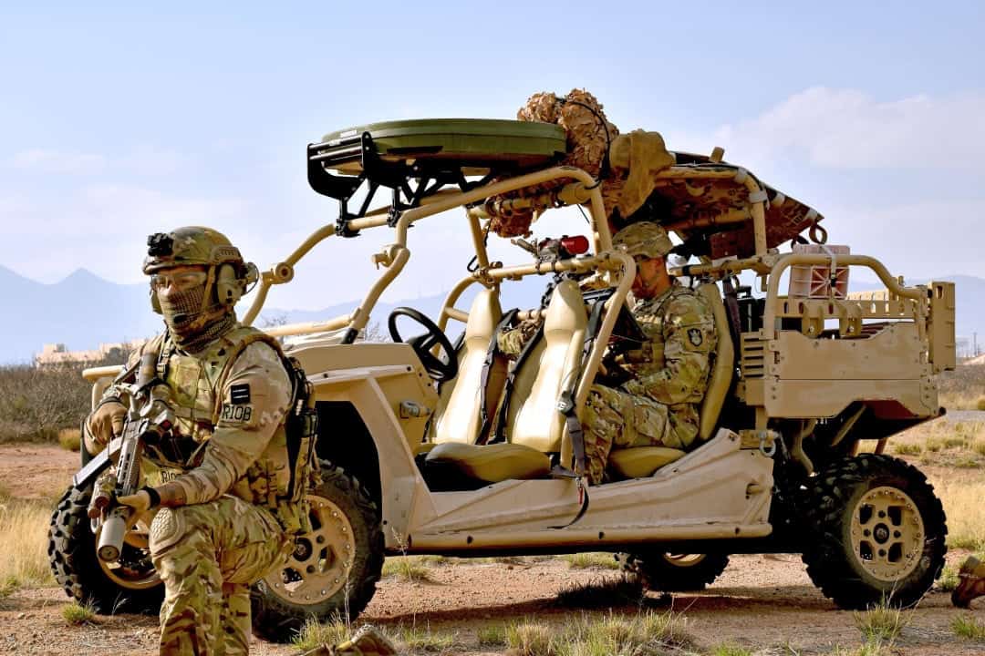 Soldiers in desert camouflage around an open-top, beige military vehicle with seats and gear in a barren field under a blue sky.